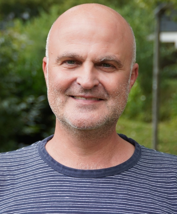 Photo of a white bald man smiling at the camera, in front of a blurred background of greenery