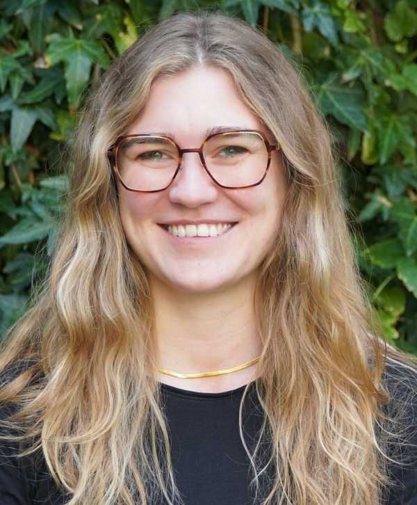 Portrait of a white woman wearing glasses and smiling at the camera, in front of a blurred background of greenery