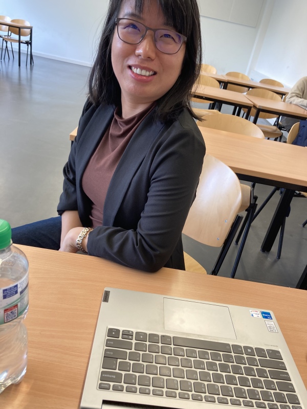 Photo of Xiaorong Gu sitting at a desk behind a laptop, smiling at the camera