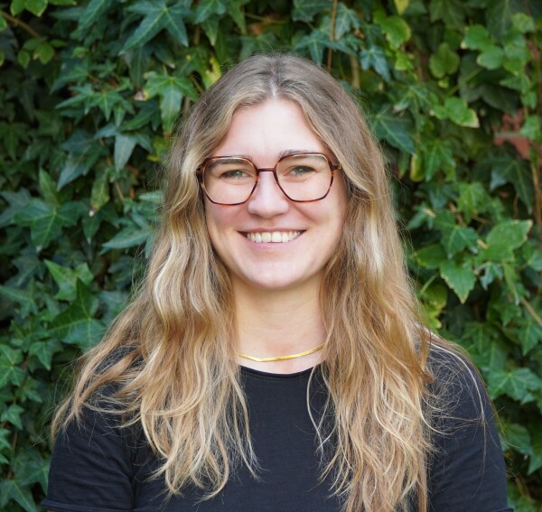 Portrait of a white woman wearing glasses and smiling at the camera, in front of a blurred background of greenery