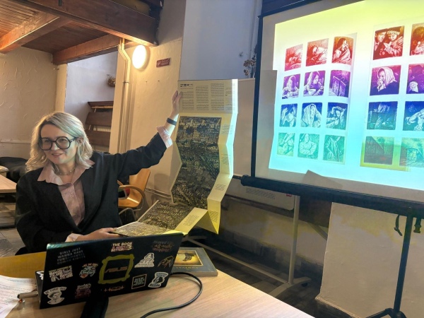 Photo of Sabina Świtała giving a presentation in front of a PowerPoint projection and holding up a long sheet of paper with text and black-and-white illustrations