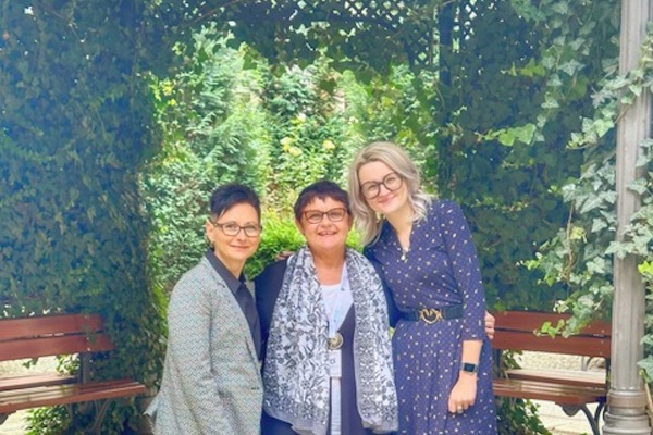 A group photo of three white woman, smiling at the camera in front of a green background