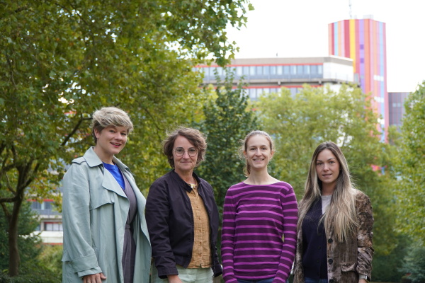 A group photo of four white woman smiling at the camera in front green trees and a colorful building